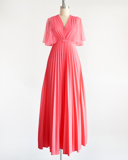 A vintage 70s coral pink maxi dress that has a faux wrap front with semi-sheer flutter sleeves, empire waist, and a long flowing accordion pleated skirt. The dress is on a dress form.