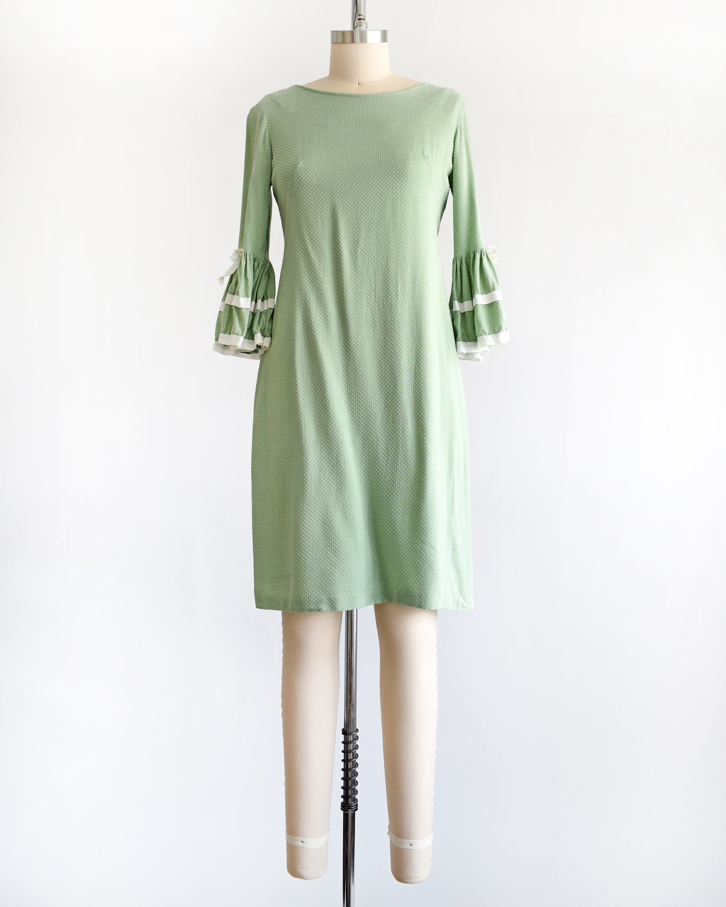 A light sage green and white Swiss dot vintage 1960s dress displayed on a dress form mannequin against a plain white background. The dress features three-quarter bell sleeves with white ruffled trim at the cuffs.