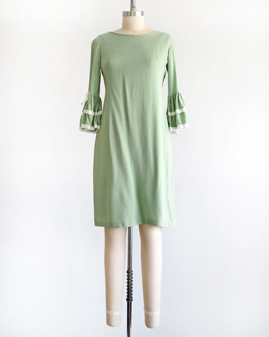 A light sage green and white Swiss dot vintage 1960s dress displayed on a dress form mannequin against a plain white background. The dress features three-quarter bell sleeves with white ruffled trim at the cuffs.