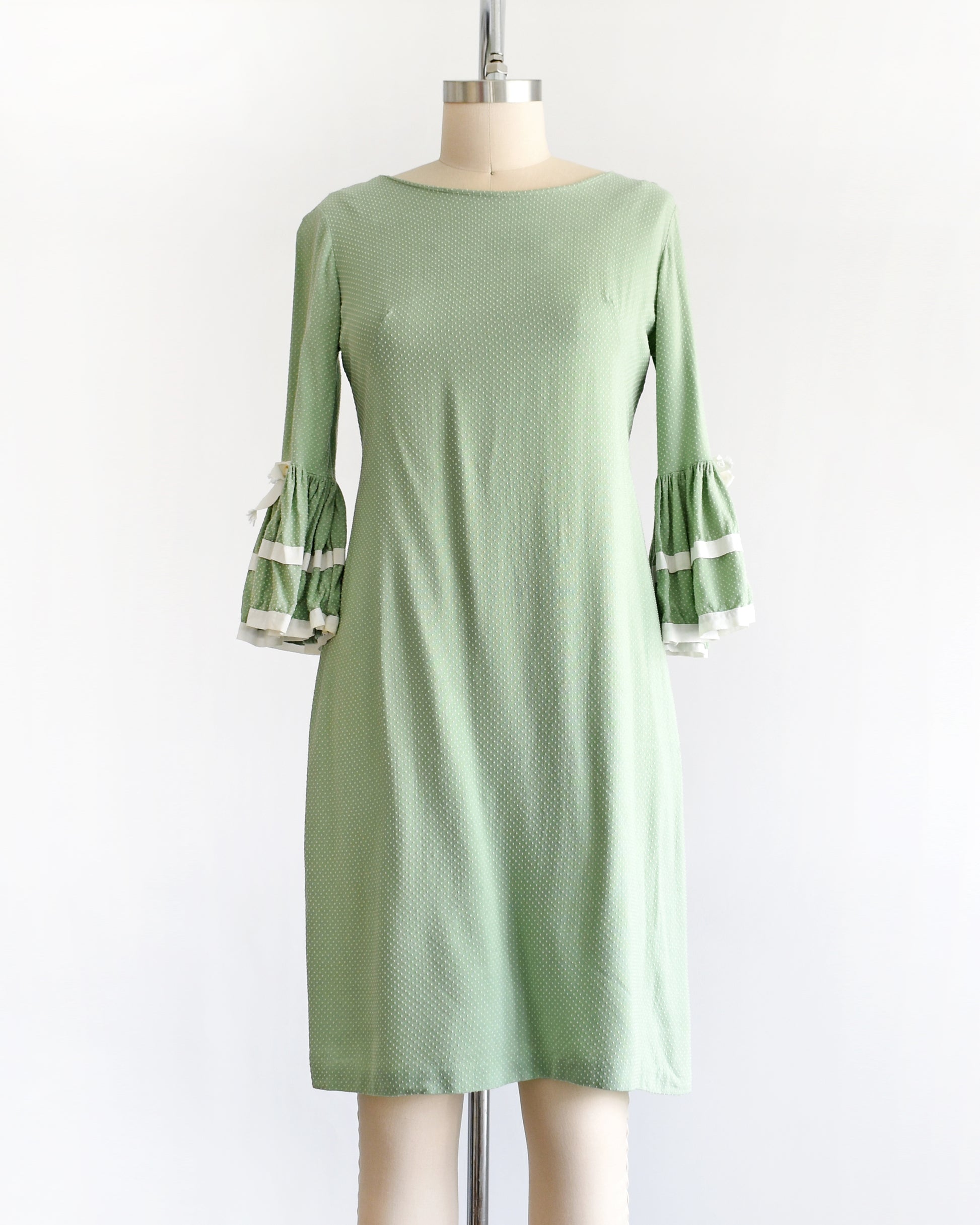 A light sage green and white Swiss dot vintage 1960s dress displayed on a dress form mannequin against a plain white background. The dress features three-quarter bell sleeves with white ruffled trim at the cuffs.
