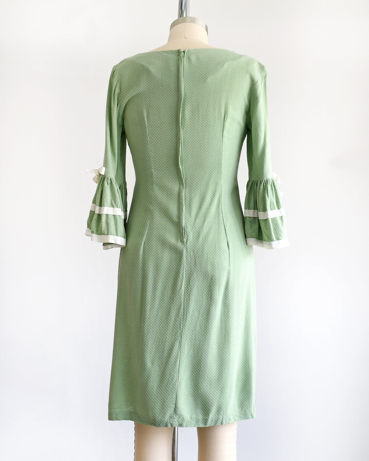 back view of a light sage green and white Swiss dot vintage 1960s dress displayed on a dress form mannequin against a plain white background. The dress features three-quarter bell sleeves with white ruffled trim at the cuffs.