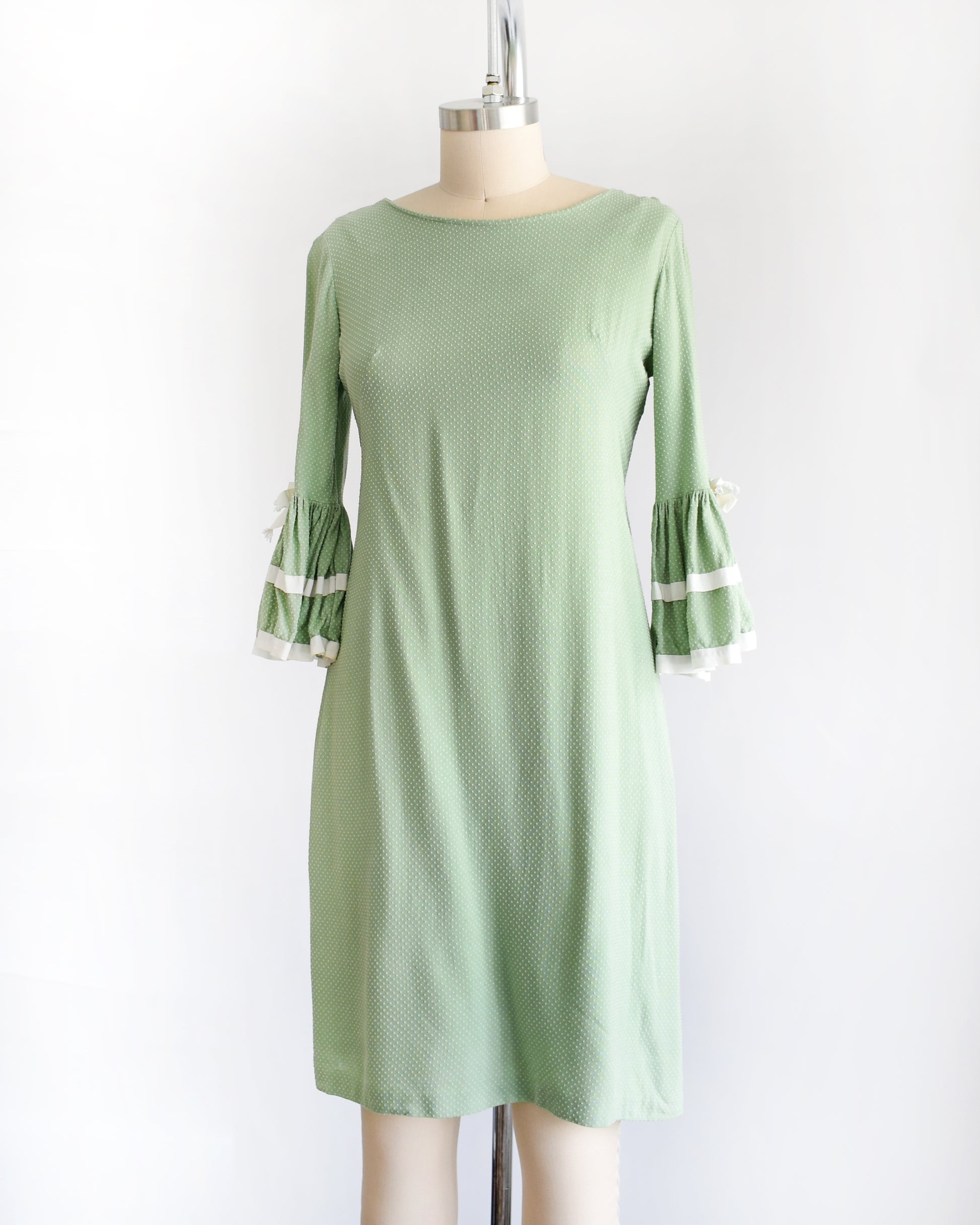 side front view of a light sage green and white Swiss dot vintage 1960s dress displayed on a dress form mannequin against a plain white background. The dress features three-quarter bell sleeves with white ruffled trim at the cuffs.