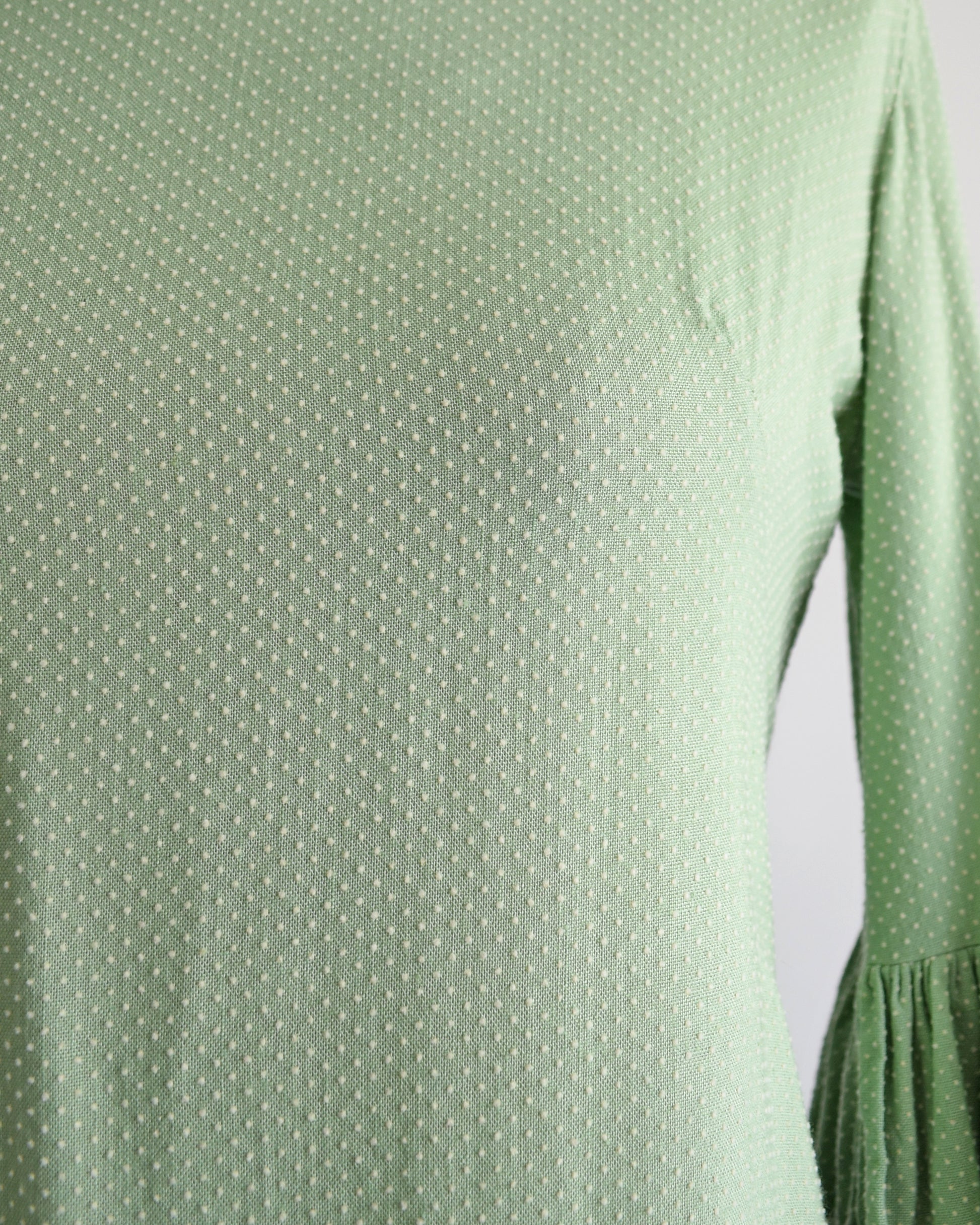 close up of the Swiss dot pattern on a light sage green vintage 1960s dress displayed on a dress form mannequin against a plain white background. The dress features three-quarter bell sleeves with white ruffled trim at the cuffs.