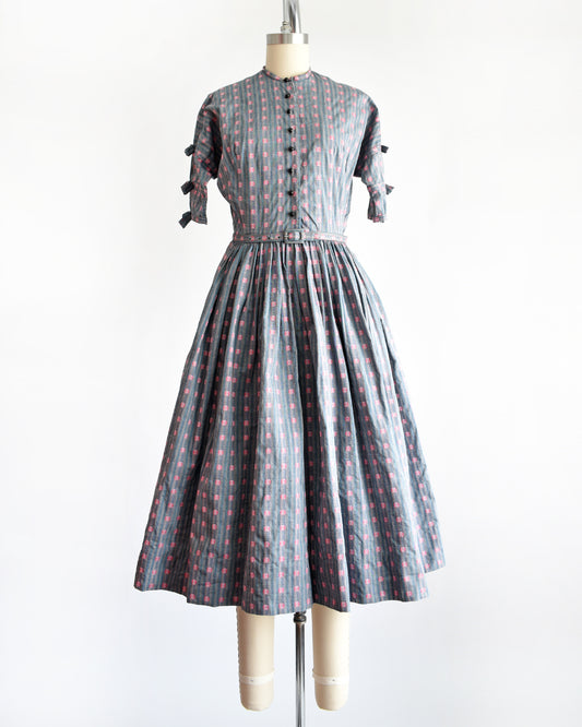 A vintage 1950s Jonathan Logan dress displayed on a mannequin against a plain white background. The dress features a gray base with a pink floral pattern, short sleeves with tie details at the cuffs, and a button-up front.