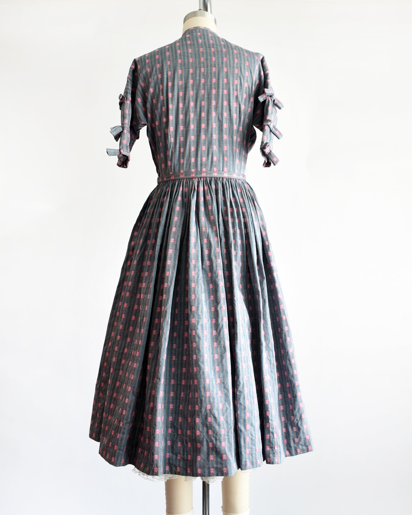 back view of a vintage 1950s Jonathan Logan dress displayed on a mannequin against a plain white background. The dress features a gray base with a pink floral pattern, short sleeves with tie details at the cuffs, and a button-up front.