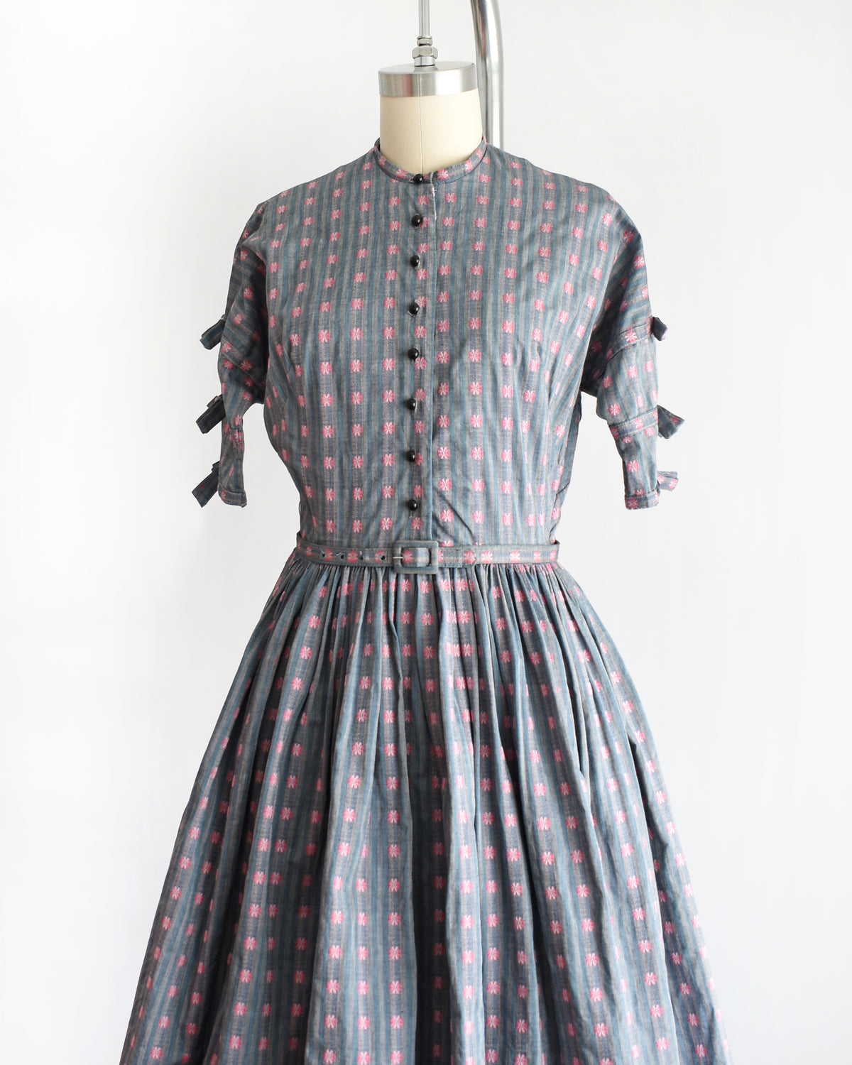 side front view of a vintage 1950s Jonathan Logan dress displayed on a mannequin against a plain white background. The dress features a gray base with a pink floral pattern, short sleeves with tie details at the cuffs, and a button-up front.