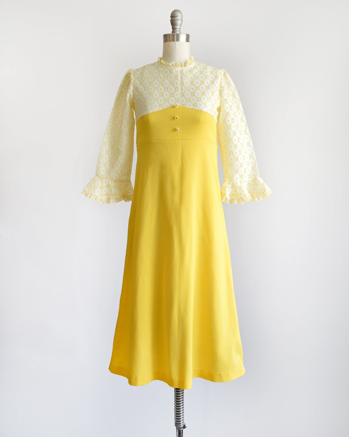 A vintage 1960s white and yellow lace mod babydoll dress with three rhinestone buttons on a dress form.