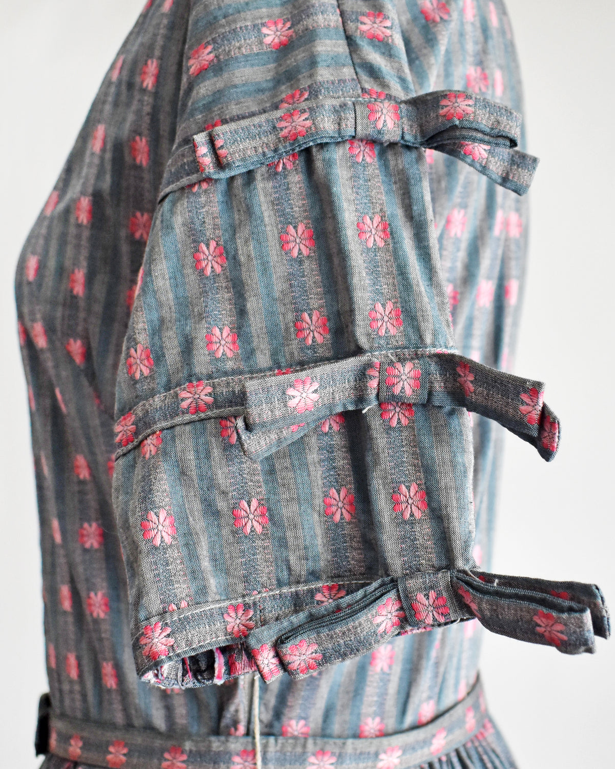 close up of the decorative bows on the sleeves of a vintage 1950s Jonathan Logan dress displayed on a mannequin against a plain white background. The dress features a gray base with a pink floral pattern, short sleeves with tie details at the cuffs, and a button-up front.
