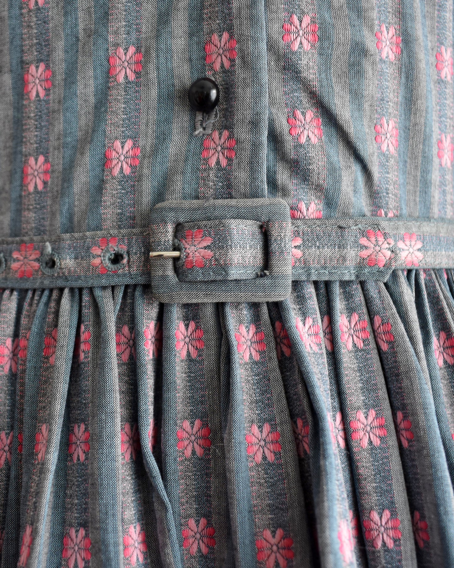 close up of the belt buckle ont the belt of a vintage 1950s Jonathan Logan dress displayed on a mannequin against a plain white background. The dress features a gray base with a pink floral pattern, short sleeves with tie details at the cuffs, and a button-up front.
