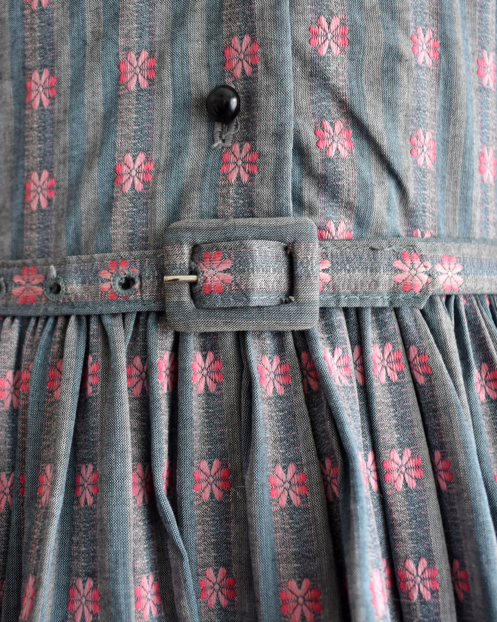 close up of the belt buckle ont the belt of a vintage 1950s Jonathan Logan dress displayed on a mannequin against a plain white background. The dress features a gray base with a pink floral pattern, short sleeves with tie details at the cuffs, and a button-up front.
