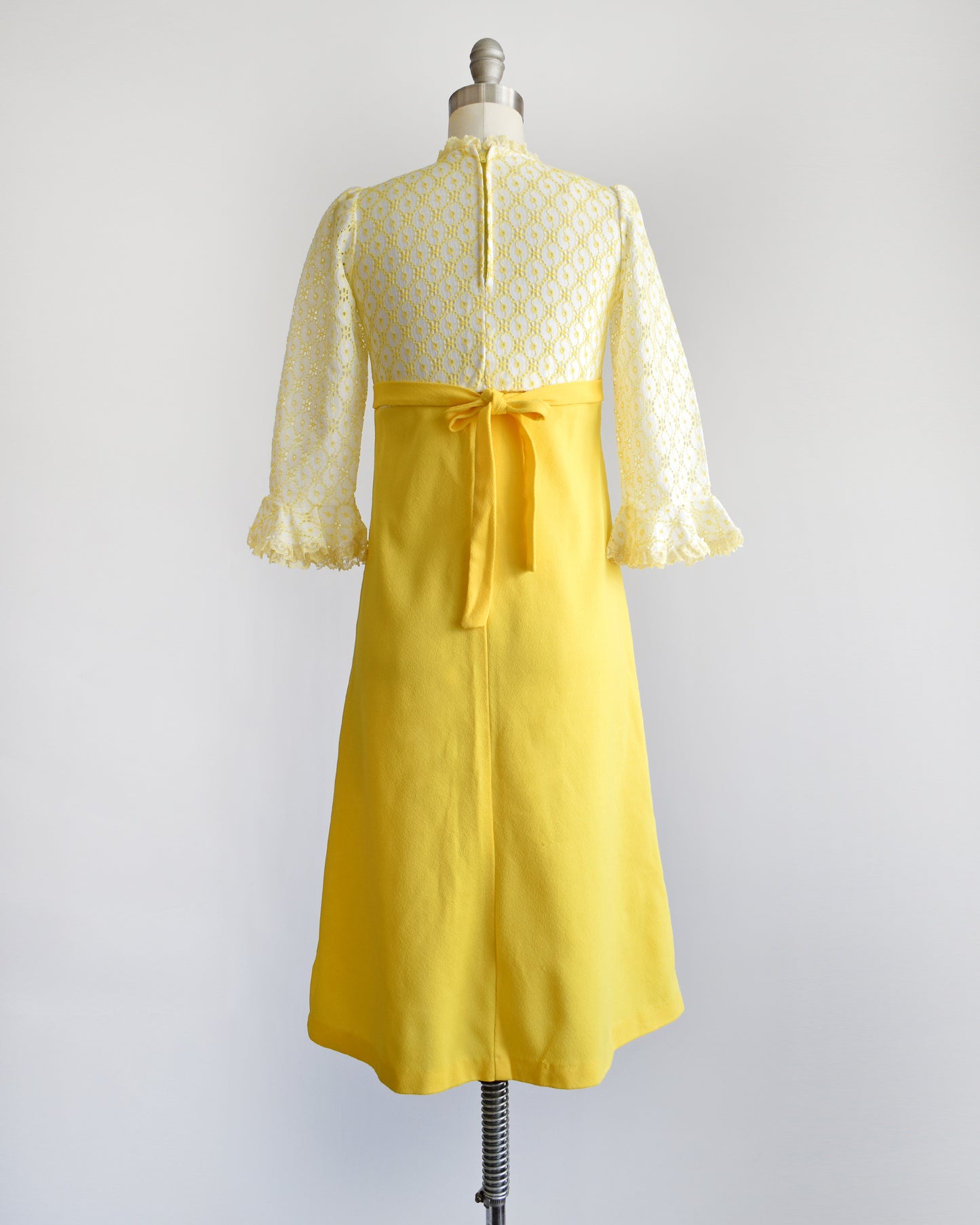 back view of vintage 1960s white and yellow lace mod babydoll dress with a tied back on a dress form.