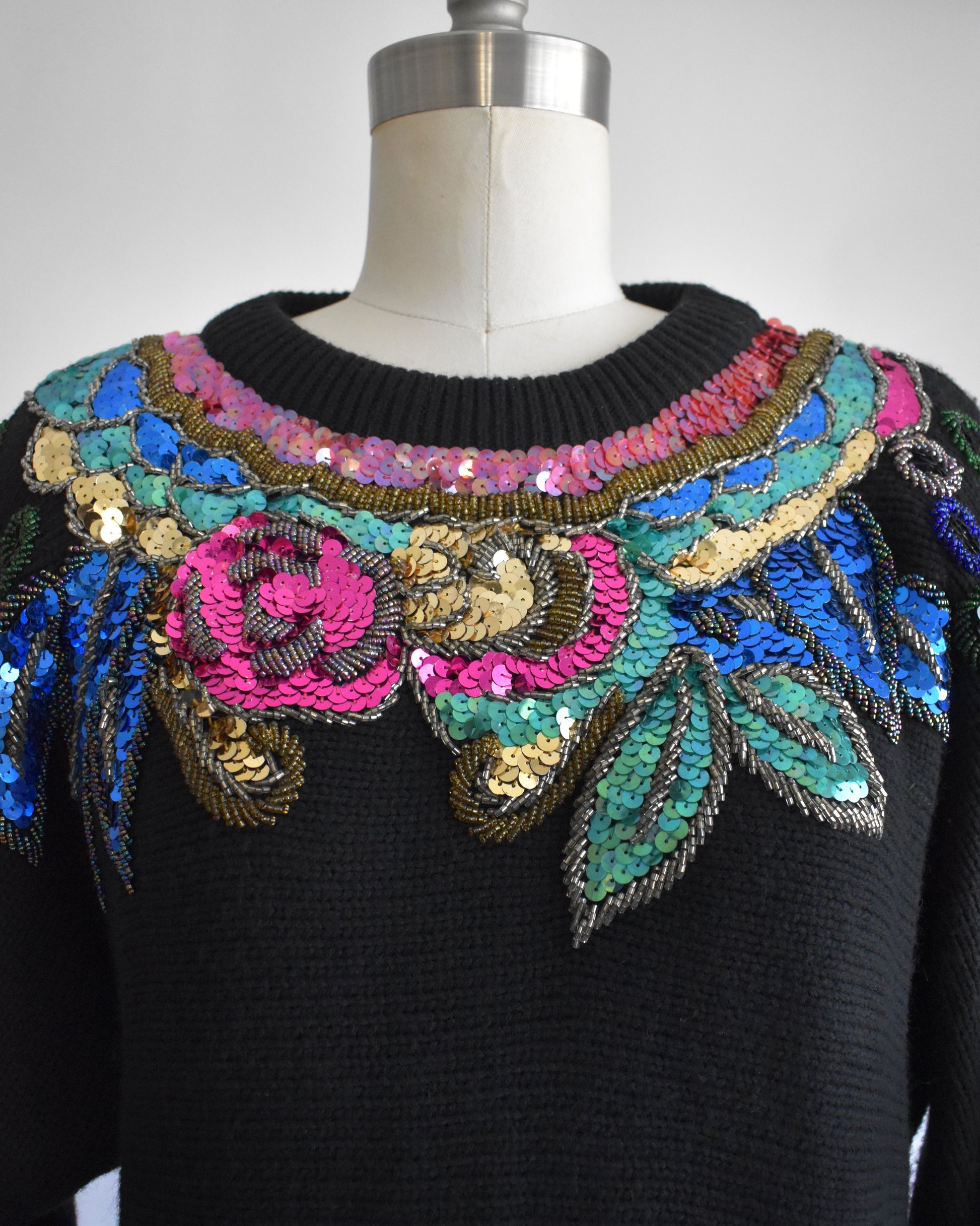 close up of the collar of a vintage 1980s black sweater with colorful floral sequin and beaded detail on a mannequin against a white background