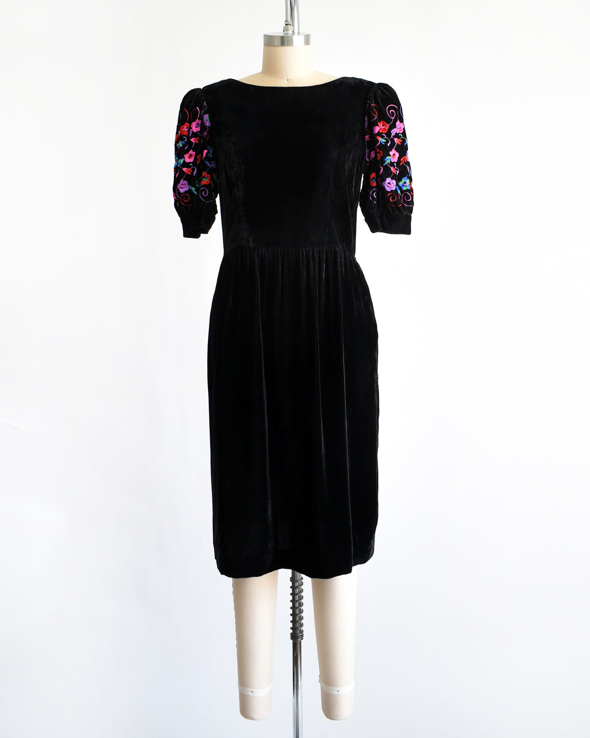 a vintage 1980s  black velvet knee-length dress by Lanz displayed on a cream dress form against a white background. The dress features short puff sleeves with colorful floral embroidery on the upper sleeves.