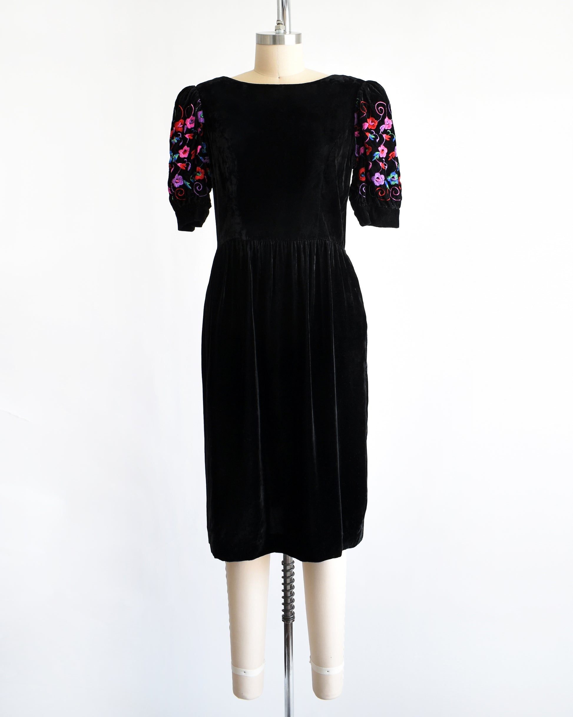 a vintage 1980s  black velvet knee-length dress by Lanz displayed on a cream dress form against a white background. The dress features short puff sleeves with colorful floral embroidery on the upper sleeves.