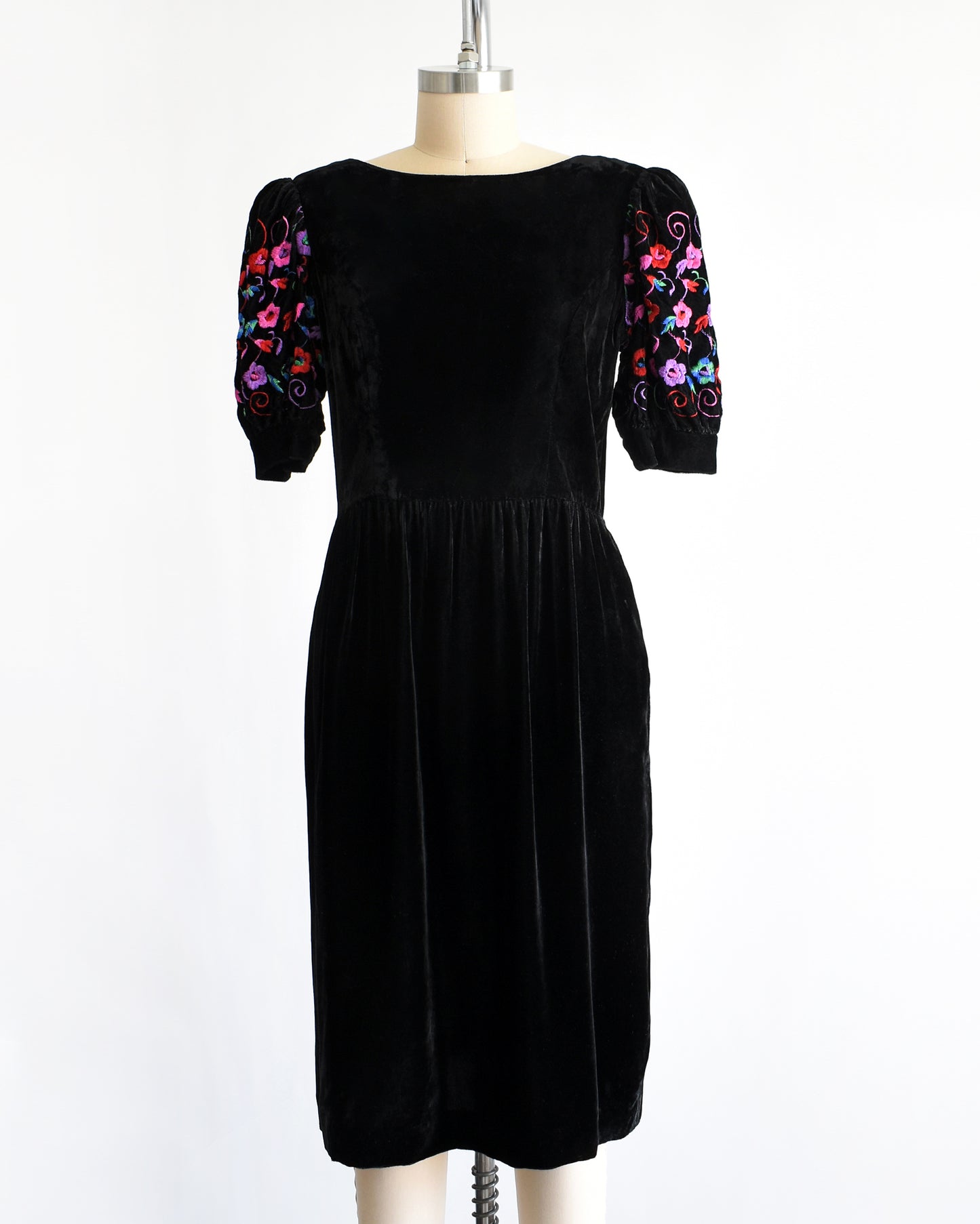 a vintage 1980s  black velvet knee-length dress by Lanz displayed on a cream dress form against a white background. The dress features short puff sleeves with colorful floral embroidery on the upper sleeves.