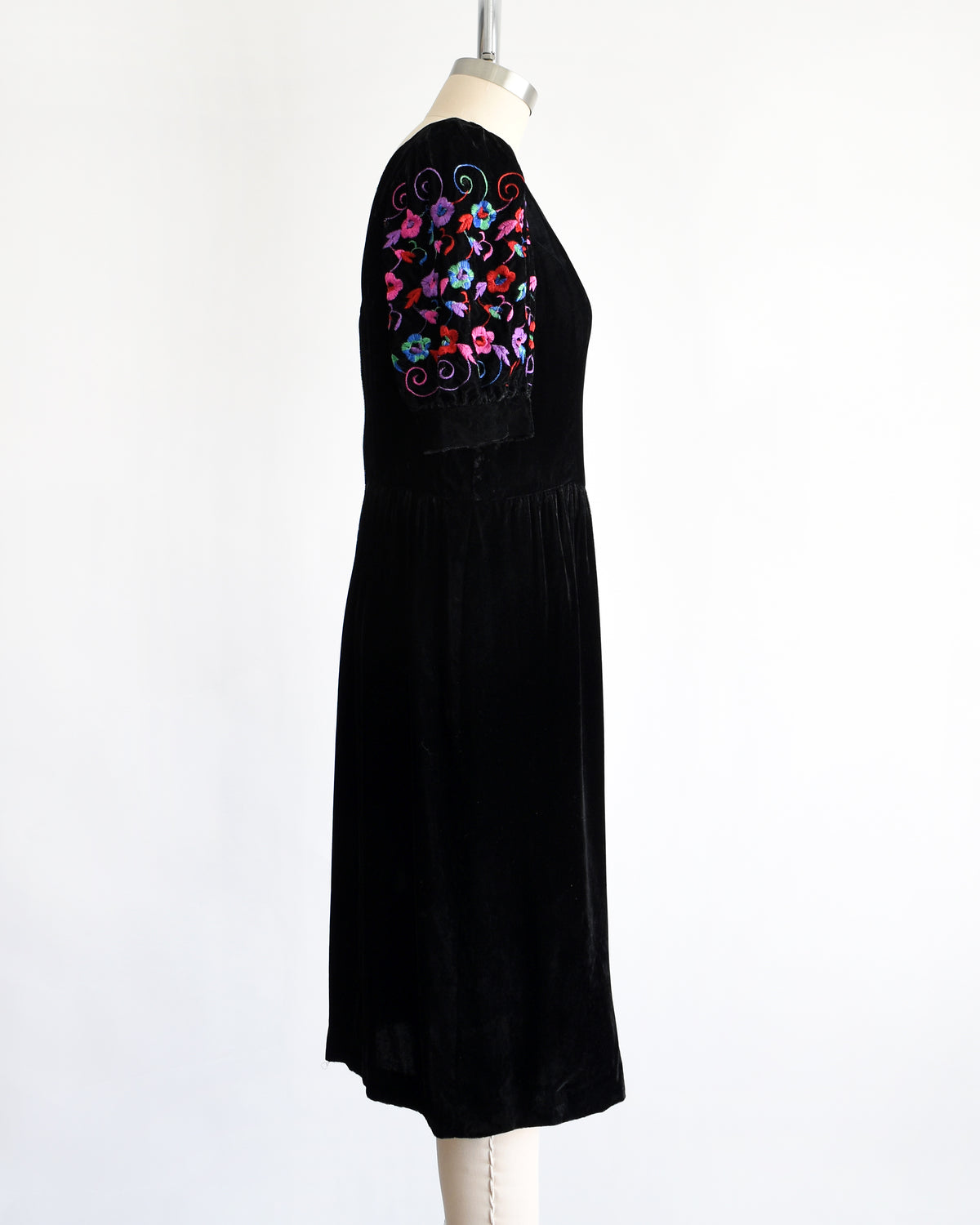 side view of a vintage 1980s  black velvet knee-length dress by Lanz displayed on a cream dress form against a white background. The dress features short puff sleeves with colorful floral embroidery on the upper sleeves.