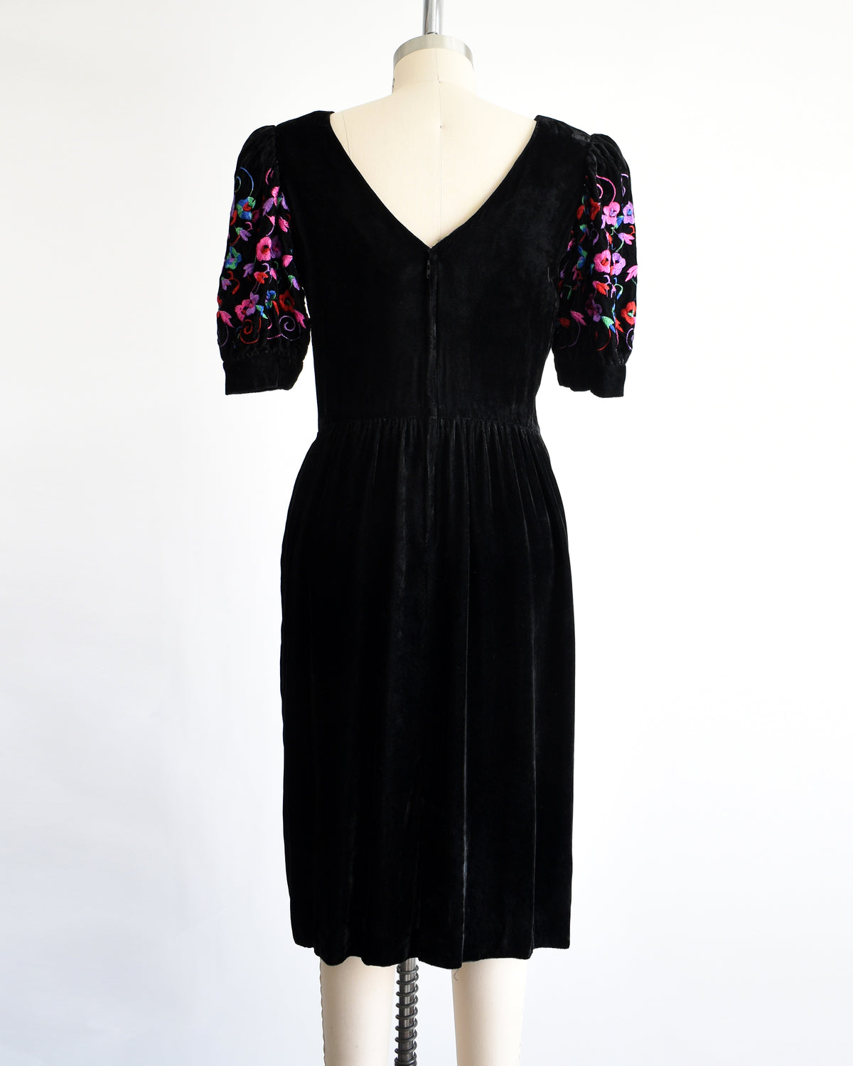 back view of a vintage 1980s  black velvet knee-length dress by Lanz displayed on a cream dress form against a white background. The dress features short puff sleeves with colorful floral embroidery on the upper sleeves.