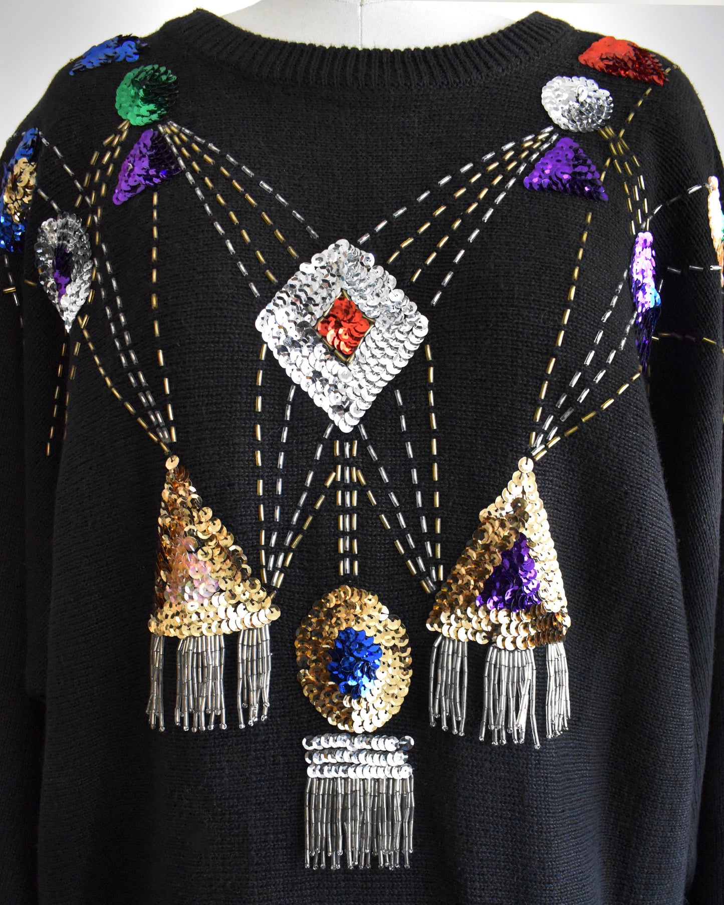 close up of a vintage black sweater with colorful gemstone sequins and tassel details on a mannequin against a white background