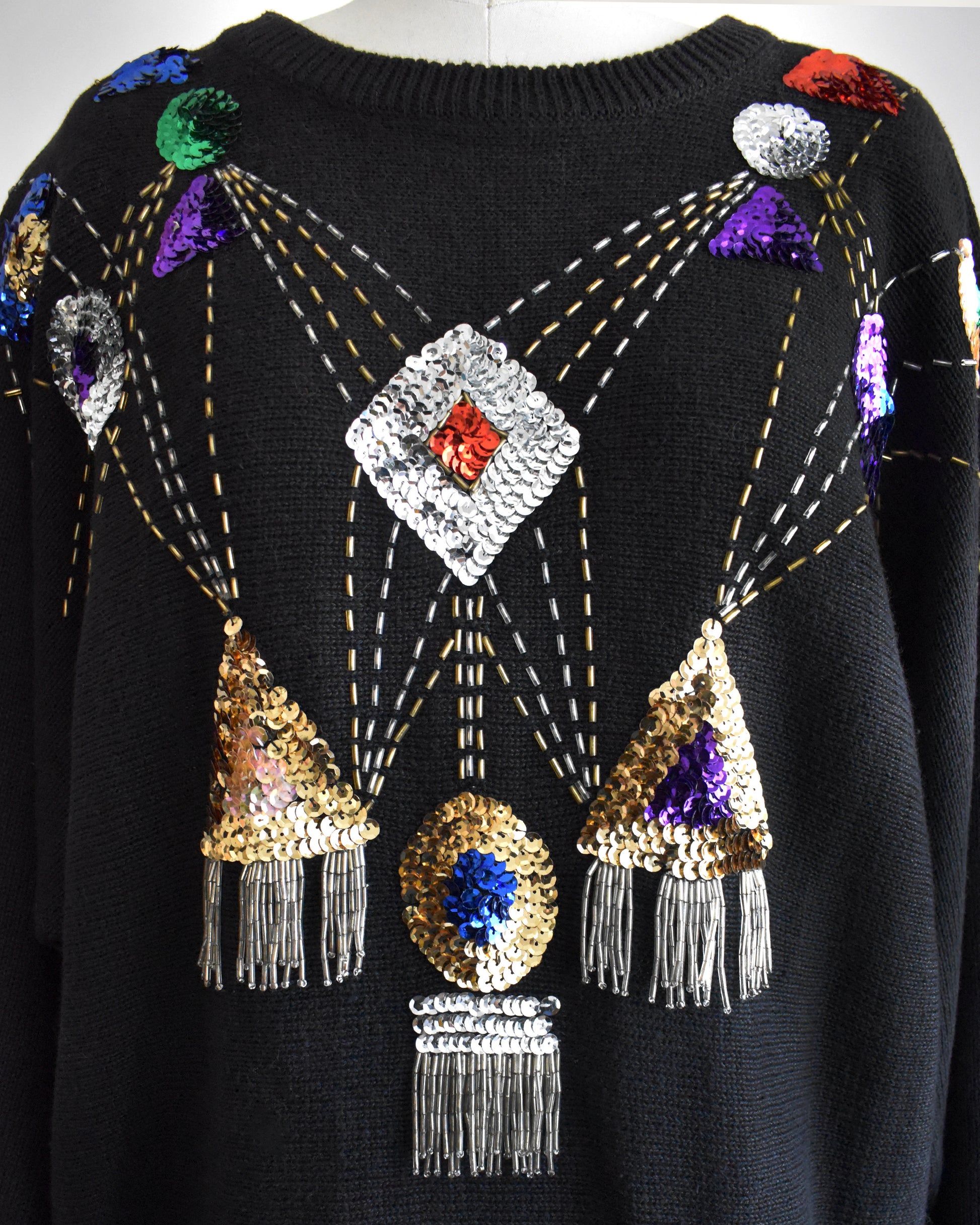 close up of a vintage black sweater with colorful gemstone sequins and tassel details on a mannequin against a white background