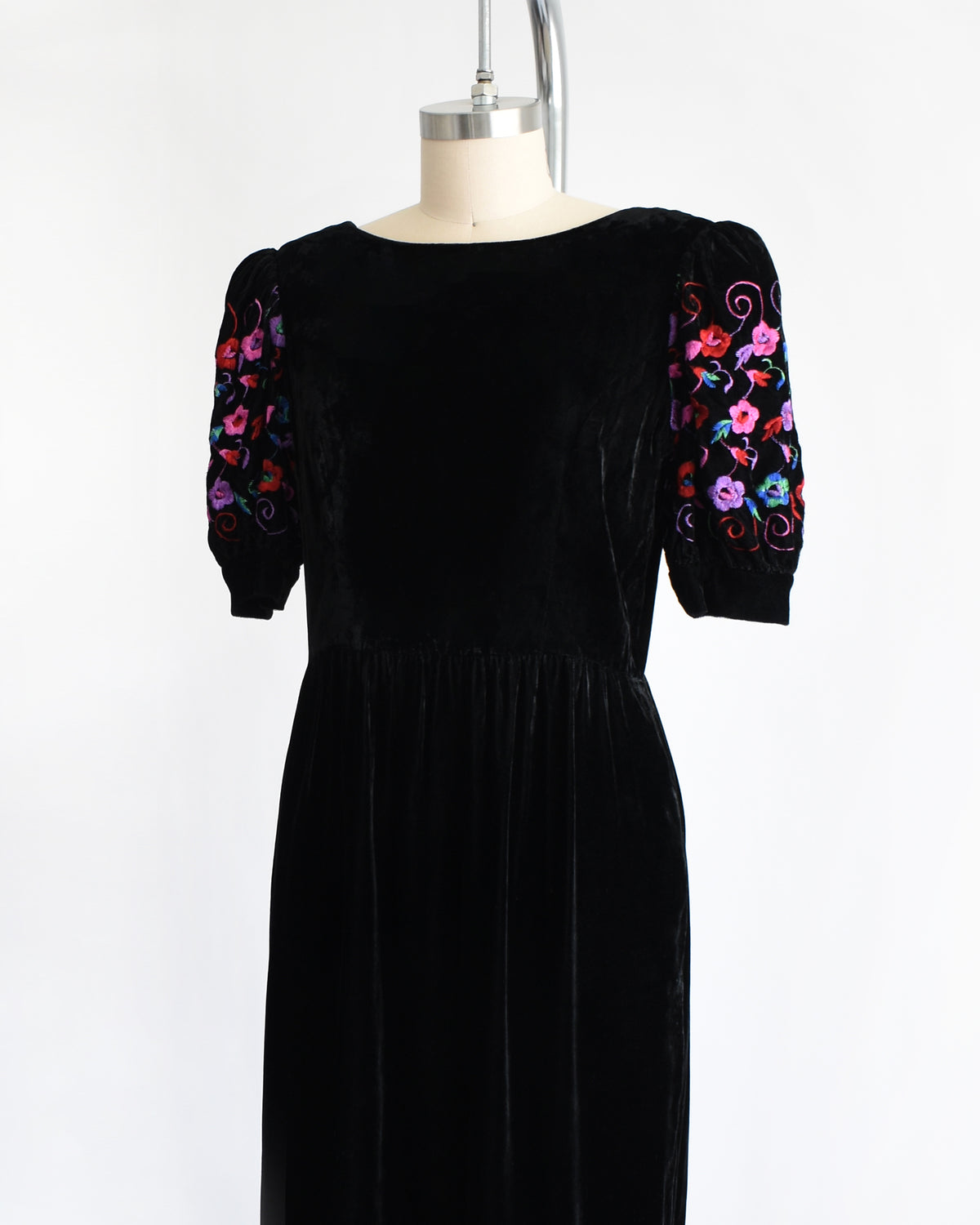 side front view of a vintage 1980s  black velvet knee-length dress by Lanz displayed on a cream dress form against a white background. The dress features short puff sleeves with colorful floral embroidery on the upper sleeves.