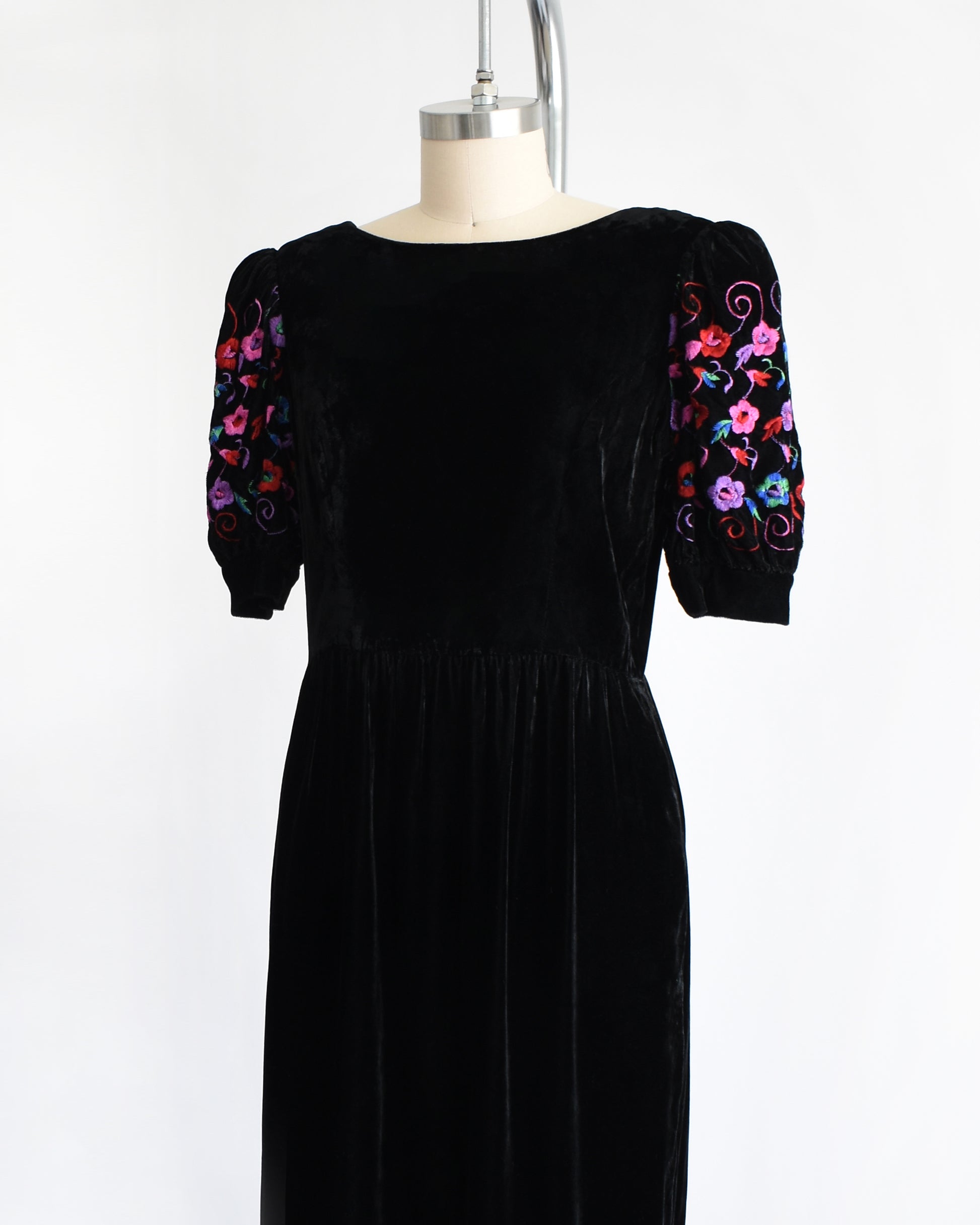 side front view of a vintage 1980s  black velvet knee-length dress by Lanz displayed on a cream dress form against a white background. The dress features short puff sleeves with colorful floral embroidery on the upper sleeves.