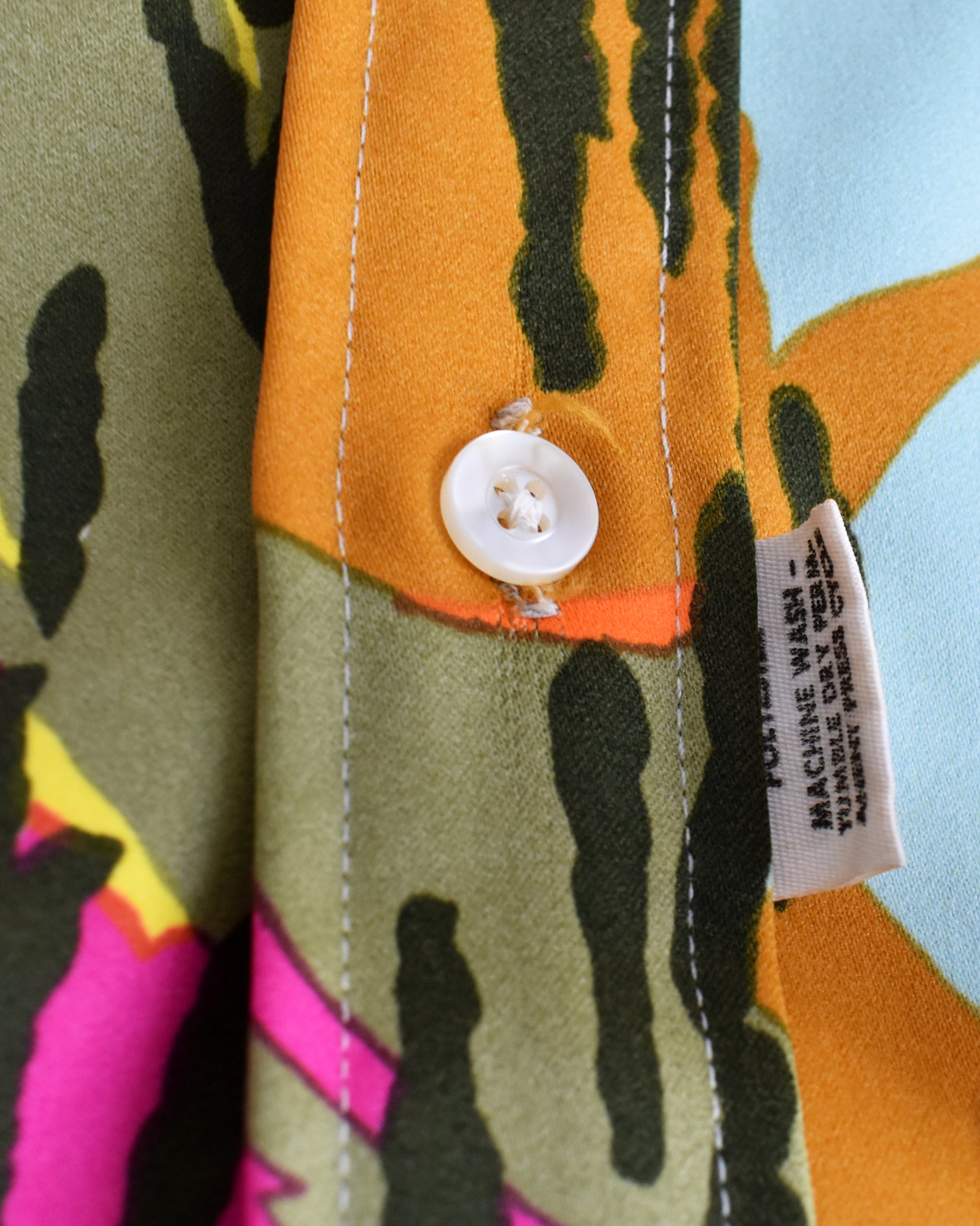 Close-up of a patterned shirt fabric with a white button and a sewn-in size/care label. The visible design combines olive green, mustard yellow, black, orange, pink, and light blue in an abstract print.