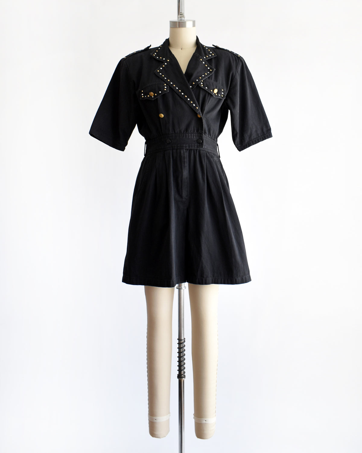 a vintage 1980s black short-sleeve romper with gold-tone hardware and studded and rhinestone detailing on the collar and chest, displayed on a cream dress form against a white background.