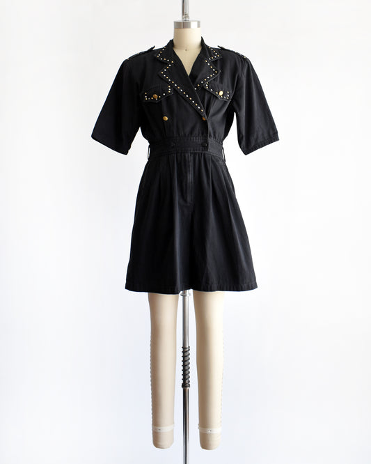 a vintage 1980s black short-sleeve romper with gold-tone hardware and studded and rhinestone detailing on the collar and chest, displayed on a cream dress form against a white background.