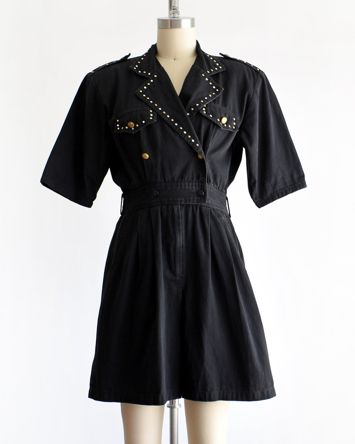 a vintage 1980s black short-sleeve romper with gold-tone hardware and studded and rhinestone detailing on the collar and chest, displayed on a cream dress form against a white background.