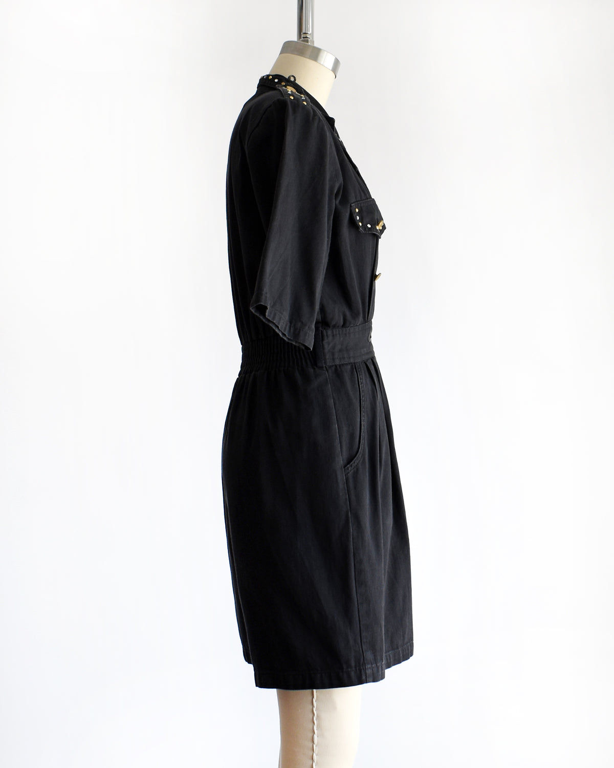 side view of a vintage 1980s black short-sleeve romper with gold-tone hardware and studded and rhinestone detailing on the collar and chest, displayed on a cream dress form against a white background.
