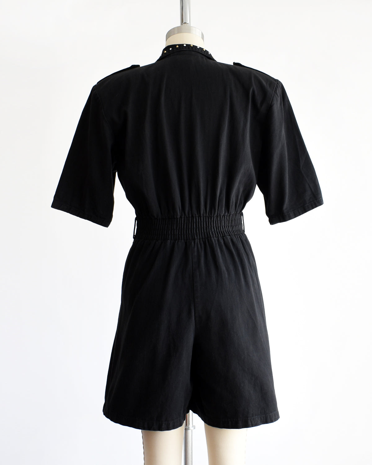 back view of a vintage 1980s black short-sleeve romper with gold-tone hardware and studded and rhinestone detailing on the collar and chest, displayed on a cream dress form against a white background.