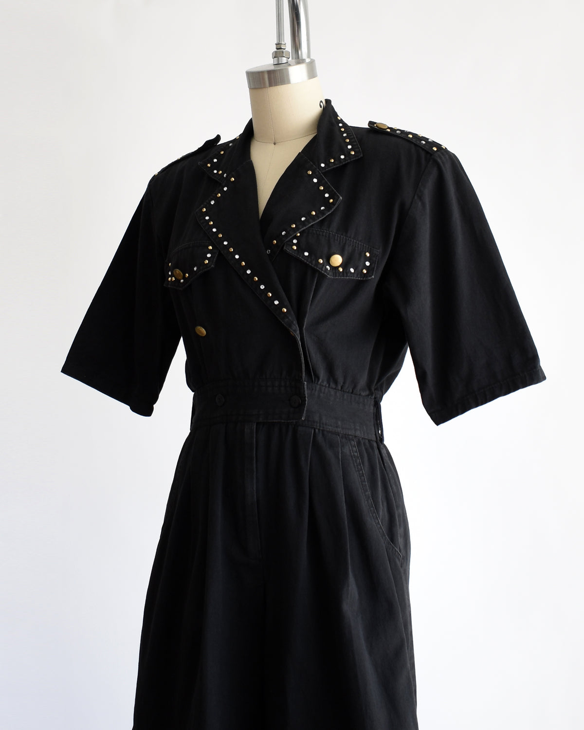 side front view of a vintage 1980s black short-sleeve romper with gold-tone hardware and studded and rhinestone detailing on the collar and chest, displayed on a cream dress form against a white background.