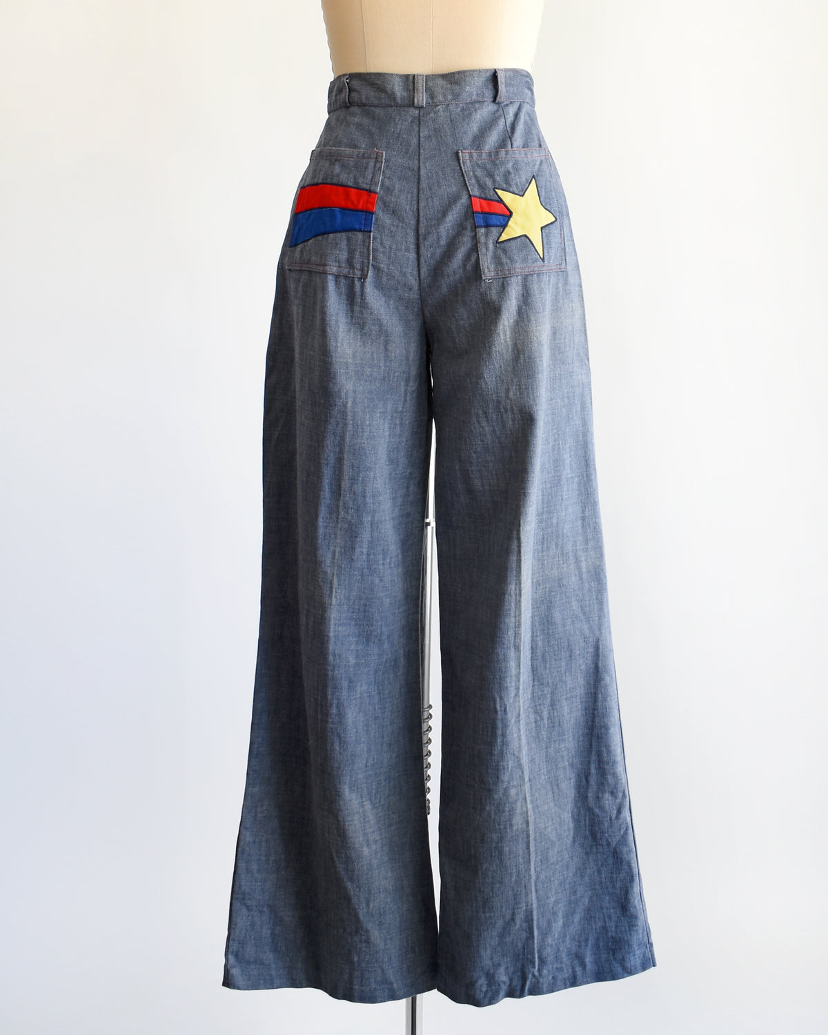 A pair of wide-leg denim pants displayed on a mannequin against a plain white background. The pants are blue denim with a yellow star patch and red and blue stripe details on the back pockets.