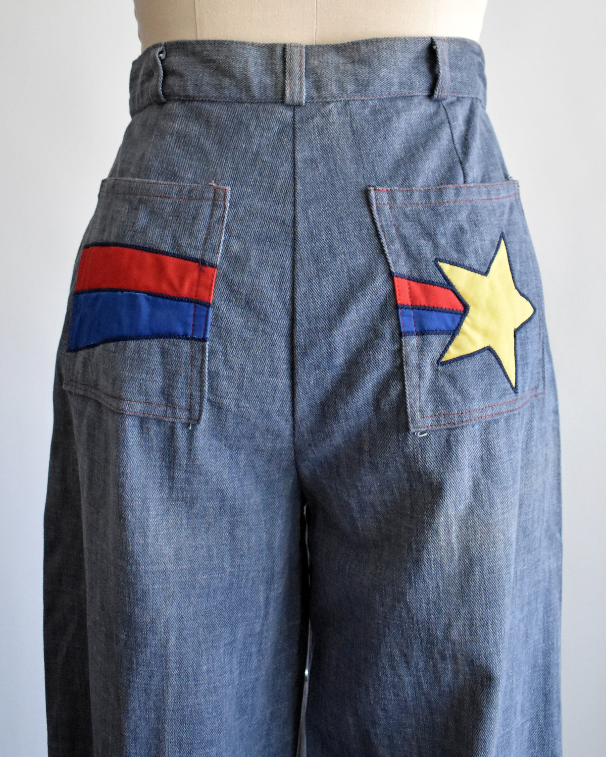 Blue denim wide-leg pants shown from the back on a mannequin, featuring a yellow star patch on one pocket and red and blue stripes on the other pocket.