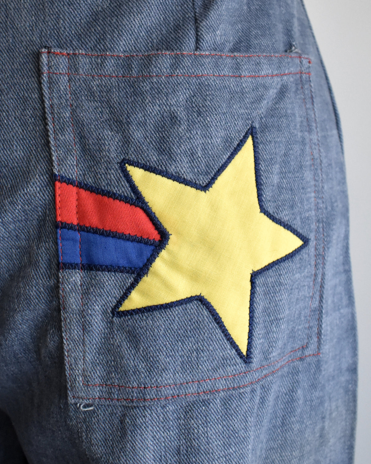 Close-up of a denim jacket pocket featuring a yellow embroidered star with a red and blue striped tail, on a blue denim background.