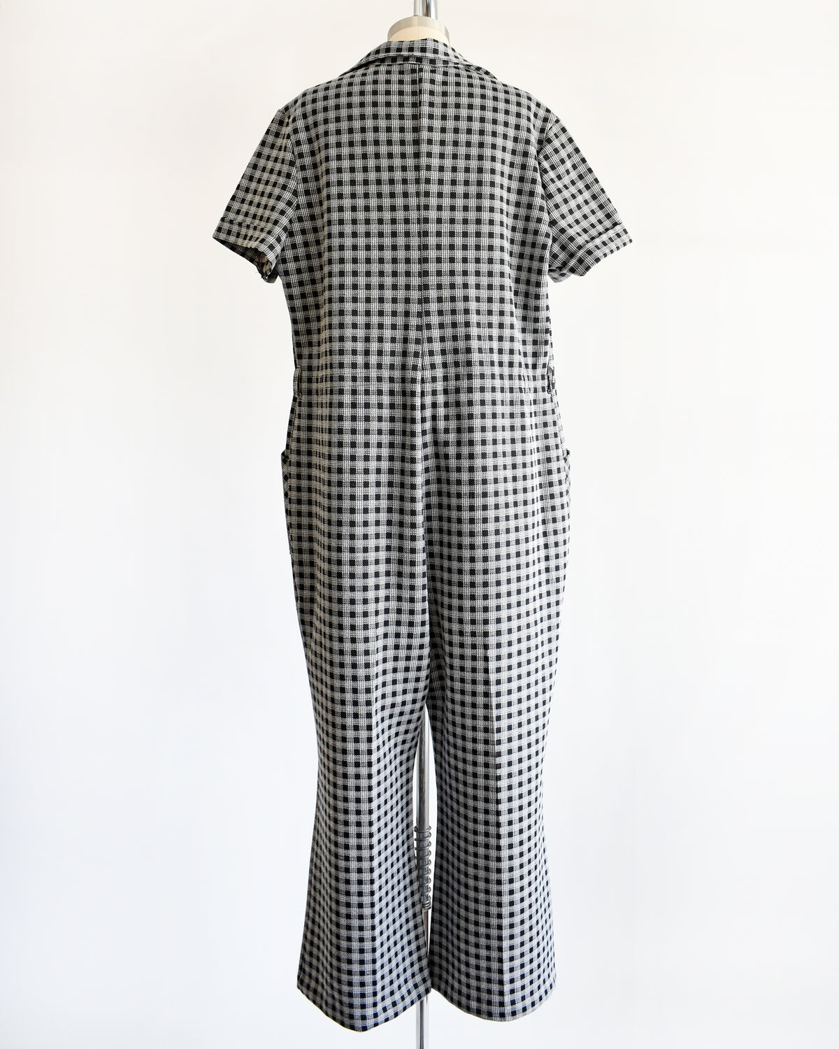 back view of a black and white gingham short-sleeve jumpsuit displayed on a dress form mannequin against a plain white background.
