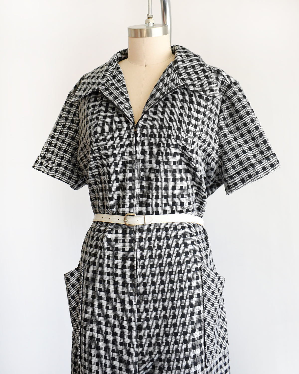 side front view of a black and white gingham short-sleeve collared shirt dress displayed on a cream dress form, styled with a thin cream belt.