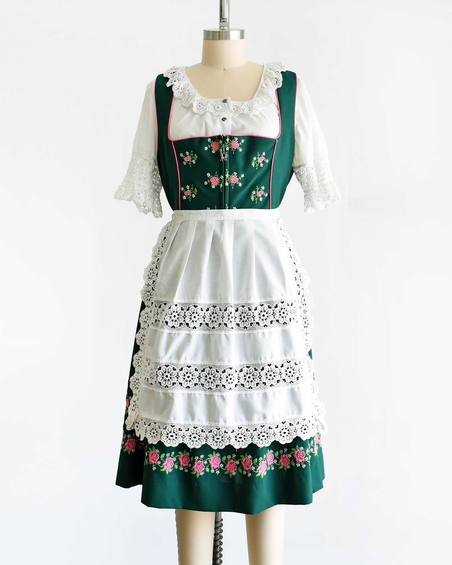 A vintage green dirndl dress that has pink and white embroidered flowers. The dress is worn with a white ruffled blouse and has a matching white apron. 