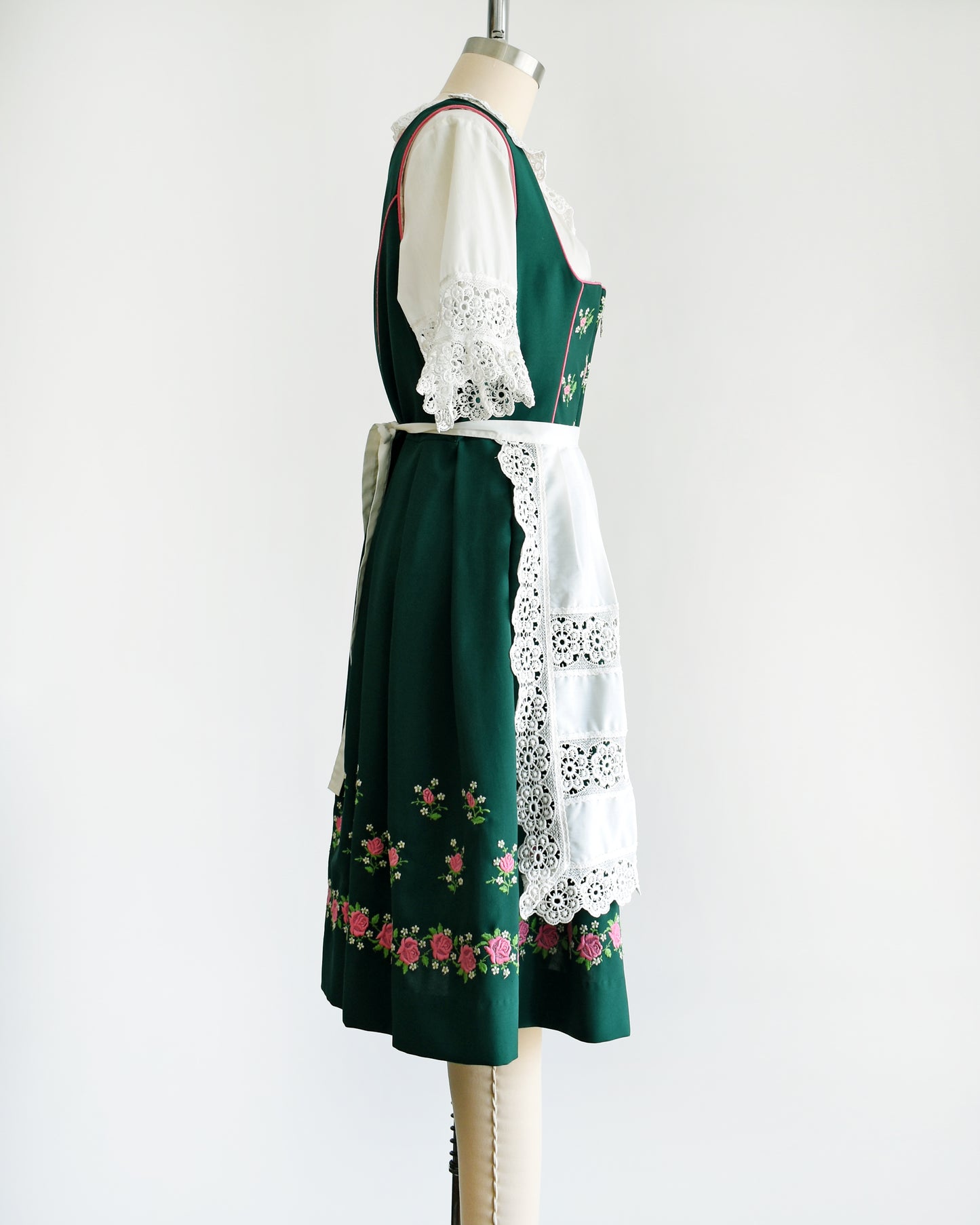 Side view of a vintage green dirndl dress that has pink and white embroidered flowers. The dress is worn with a white ruffled blouse and has a matching white apron. 