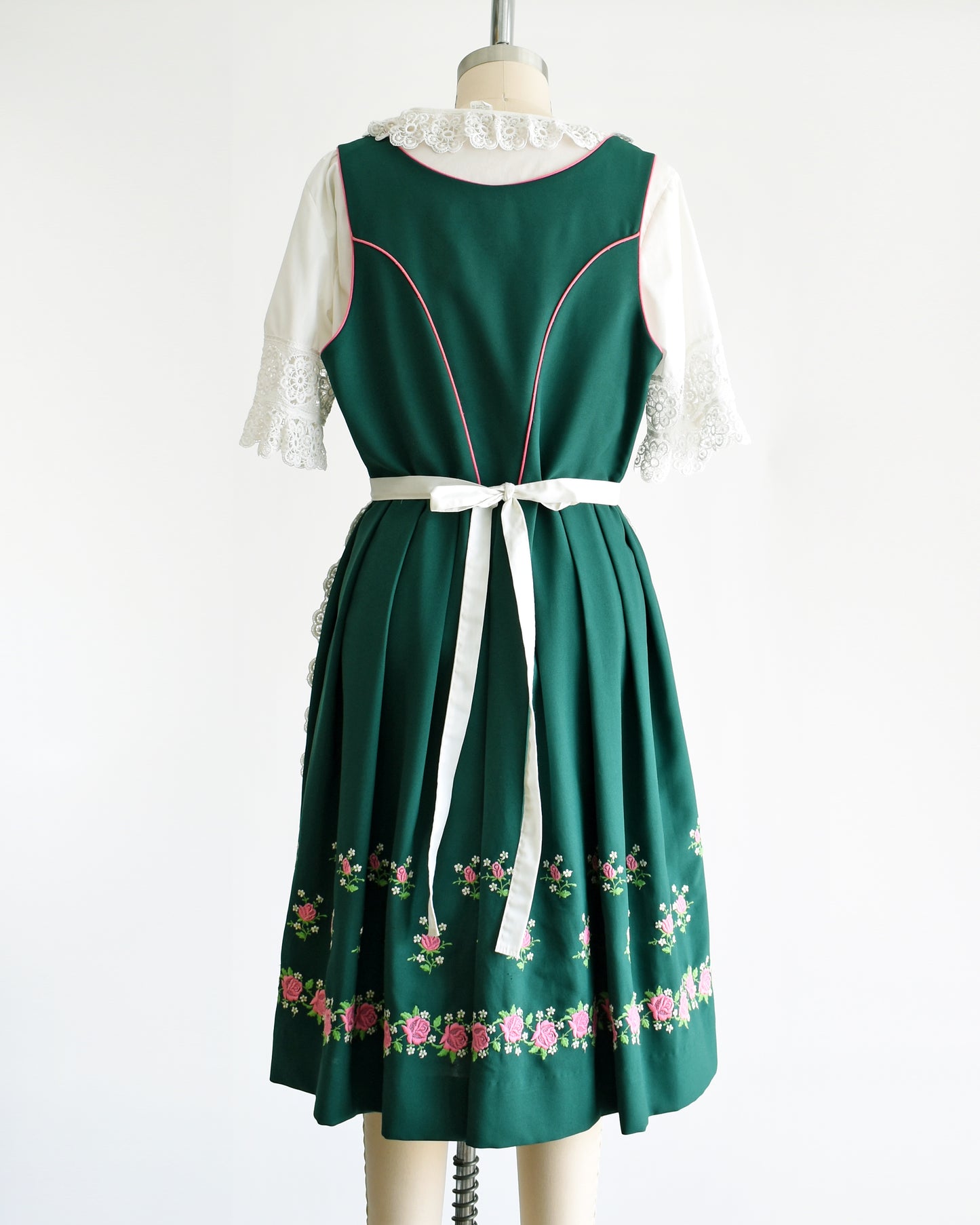 Back view of a vintage green dirndl dress that has pink and white embroidered flowers. The dress is worn with a white ruffled blouse and has a matching white apron. 