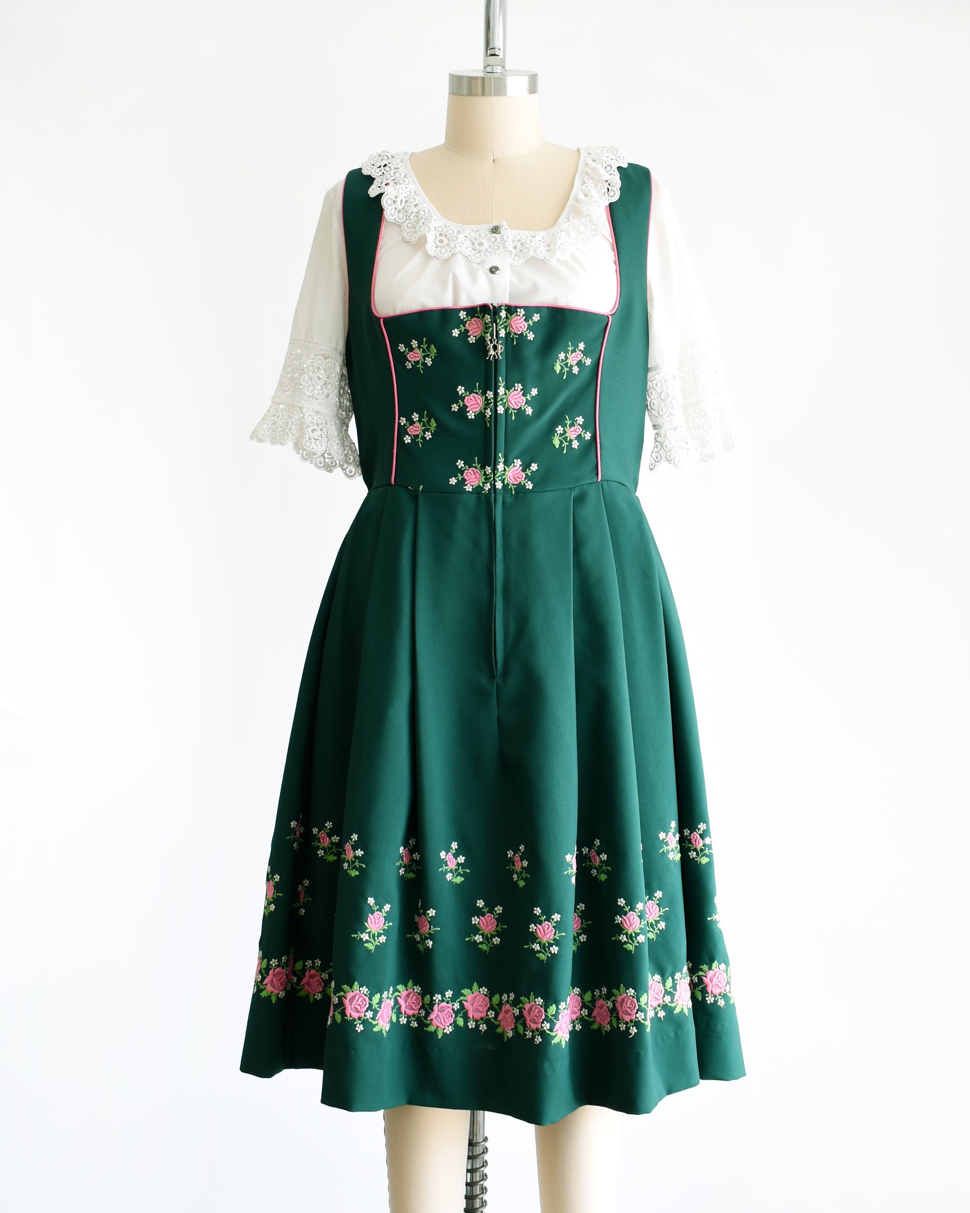 A vintage green dirndl dress that has pink and white embroidered flowers. The dress is worn with a white ruffled blouse. The dress is shown without the apron