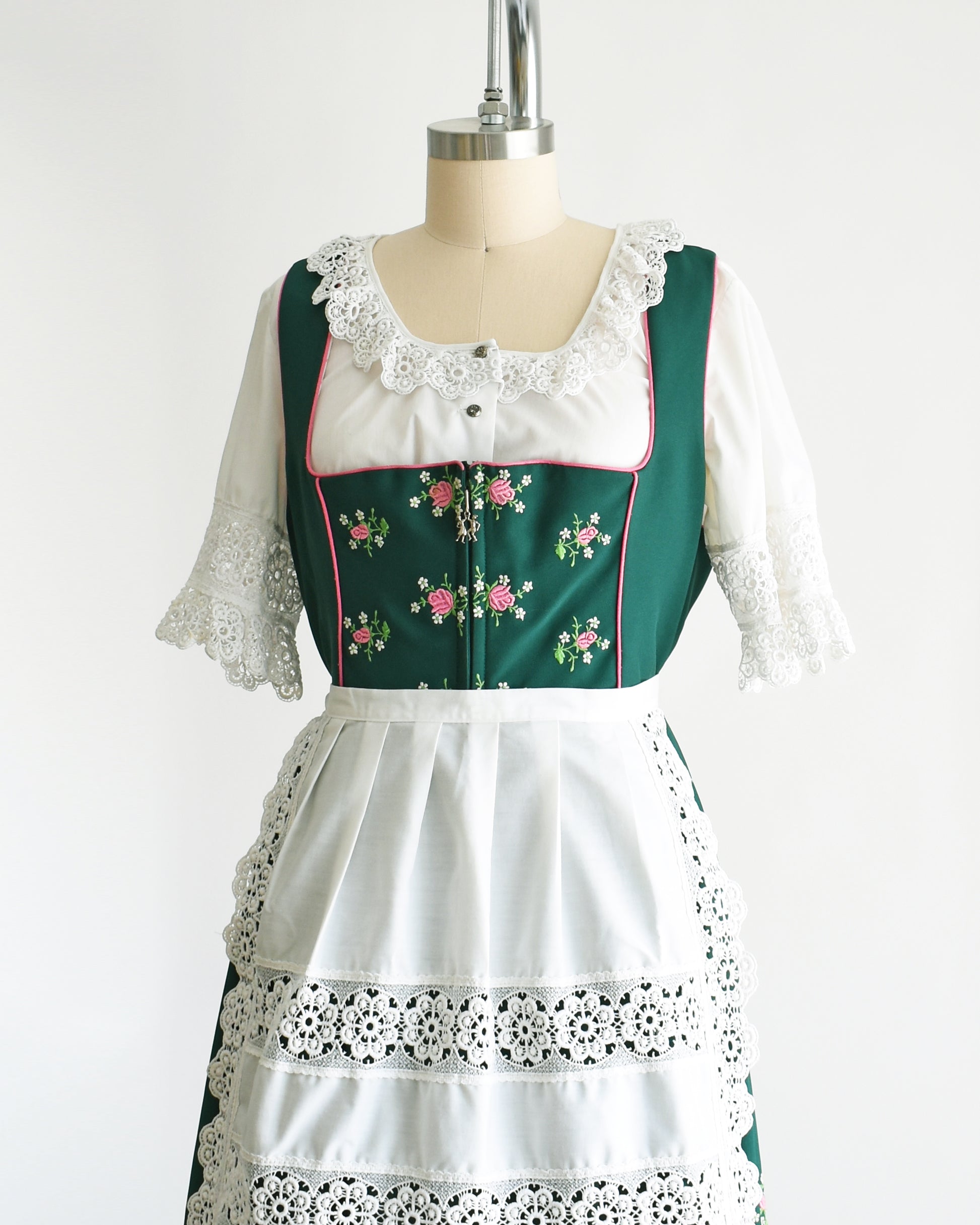 Side front view of a vintage green dirndl dress that has pink and white embroidered flowers. The dress is worn with a white ruffled blouse and has a matching white apron. 