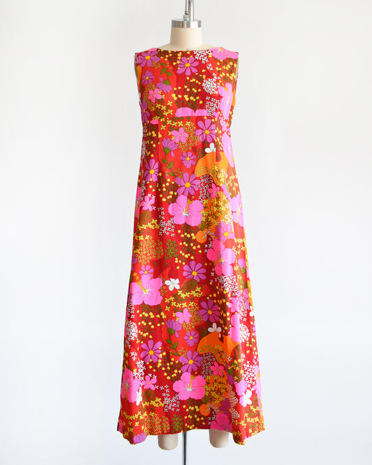 A vintage 1960s Ui-Maikai Hawaiian maxi dress features a bright and bold vibrant floral pattern set on a combination of deep and bright red