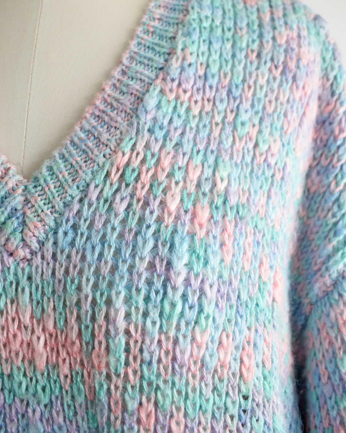 Close-up of a hand-knit V-neck sweater in variegated pastel yarn with pink, mint green, and lavender tones, displayed on a cream-colored dress form.
