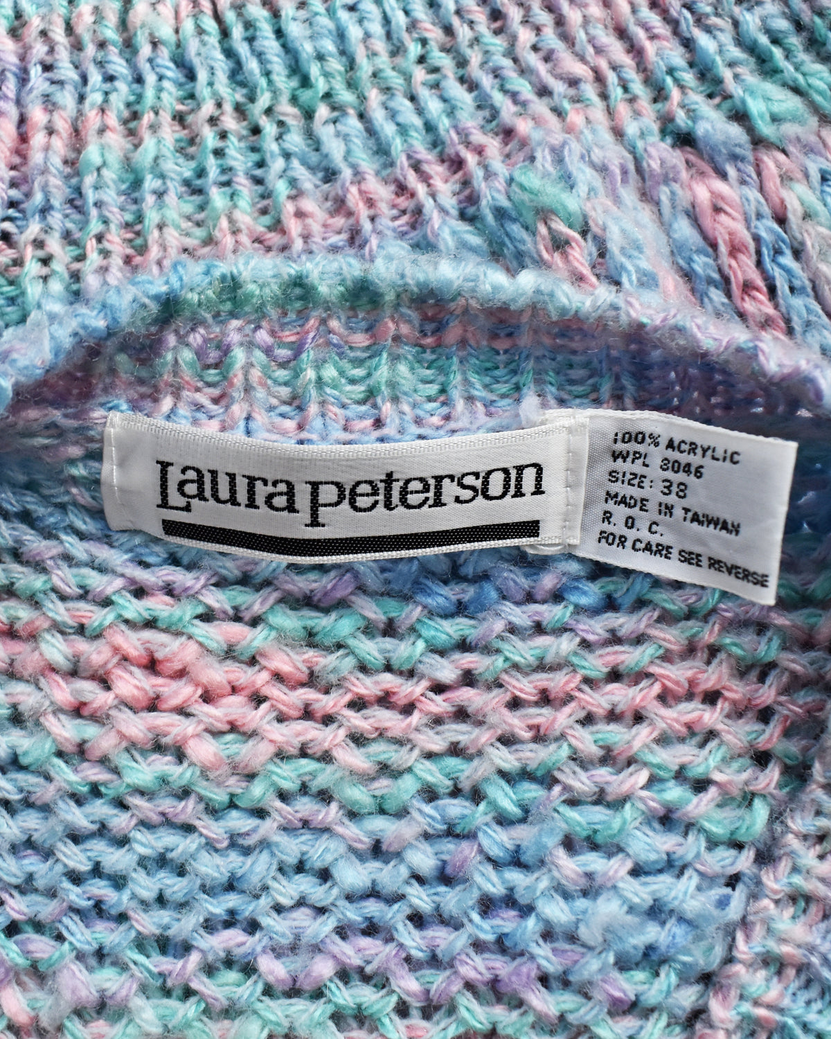 Close-up of a knitted sweater with a visible Laura Peterson brand label, showing a variegated pastel color mix of pink, blue, and mint in a textured knit pattern.