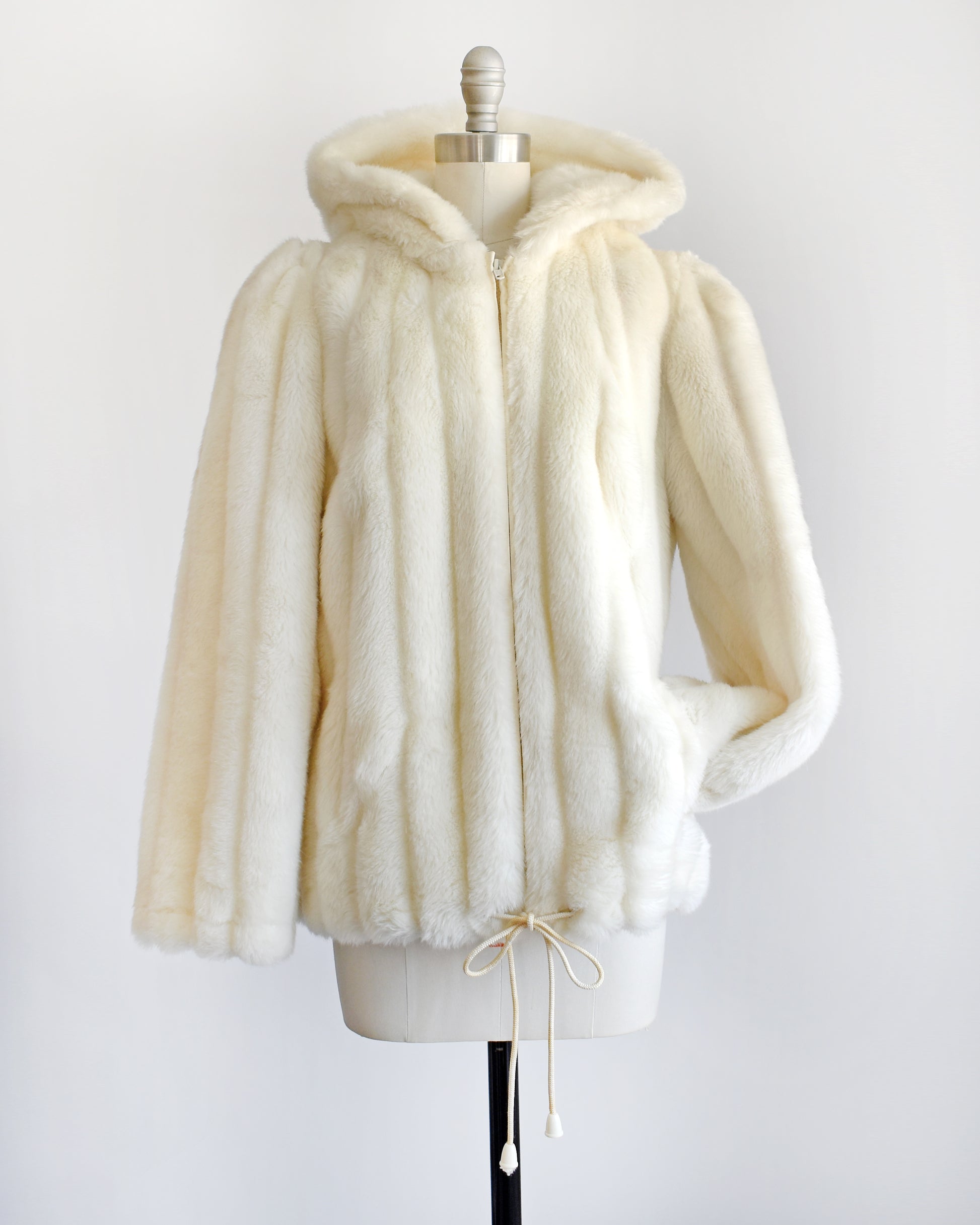 a vintage 1980s cream faux fur hooded coat with a drawstring on the bottom. a sleeve is tucked into one of the pockets on the coat.