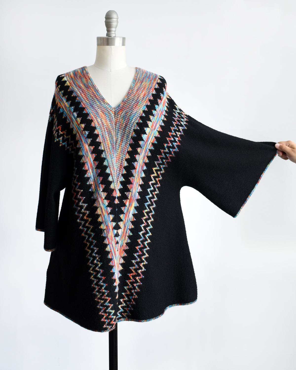 a vintage 1970s  black three-quarter sleeve tunic top with a colorful geometric zigzag pattern down the front, displayed on a mannequin against a white background with a hand holding out one sleeve against a plain white background.