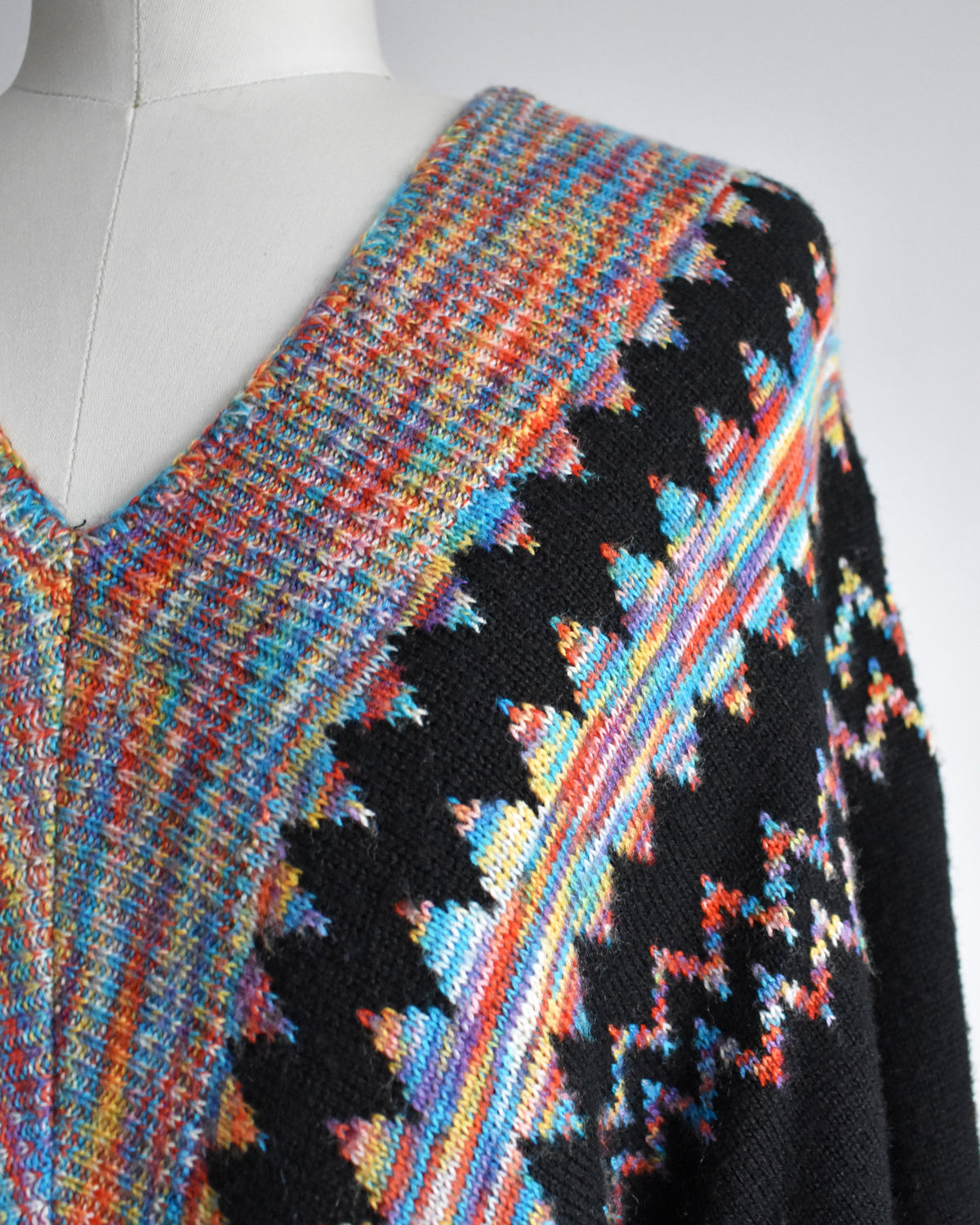 A close-up of a knit sweater on a mannequin, featuring a black base with a colorful geometric pattern and a striped multicolor yoke.
