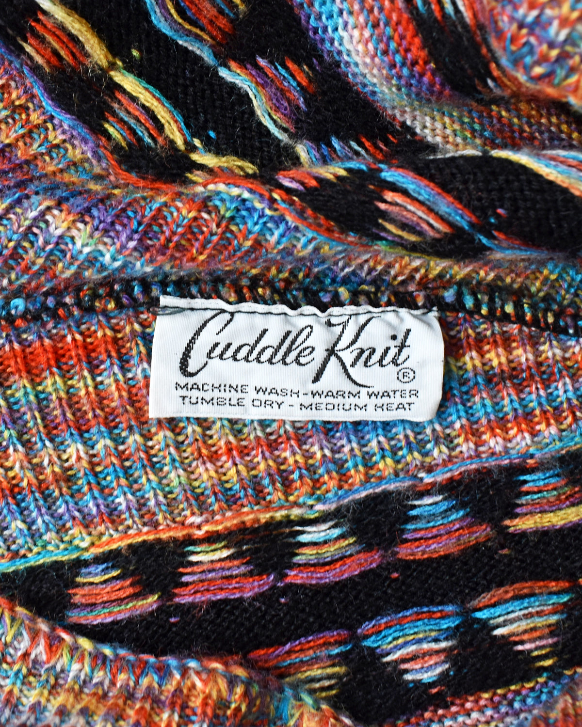 Close-up of a multicolor knitted garment with a white sewn-in label reading 'Cuddle Knit' and care instructions.