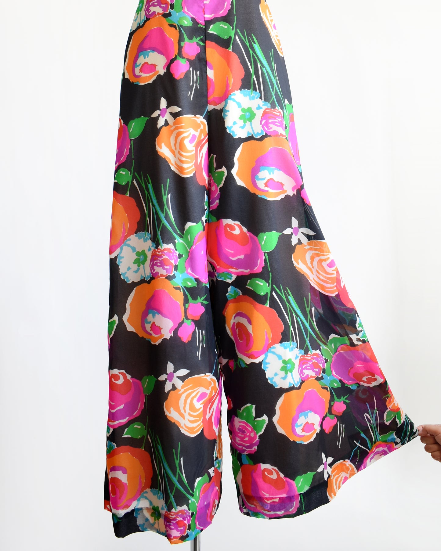 the bottom half of a vintage 1970s black floral print jumpsuit by Malia on a dress form. One of the pant legs is being pulled to show the width of the pant hem.